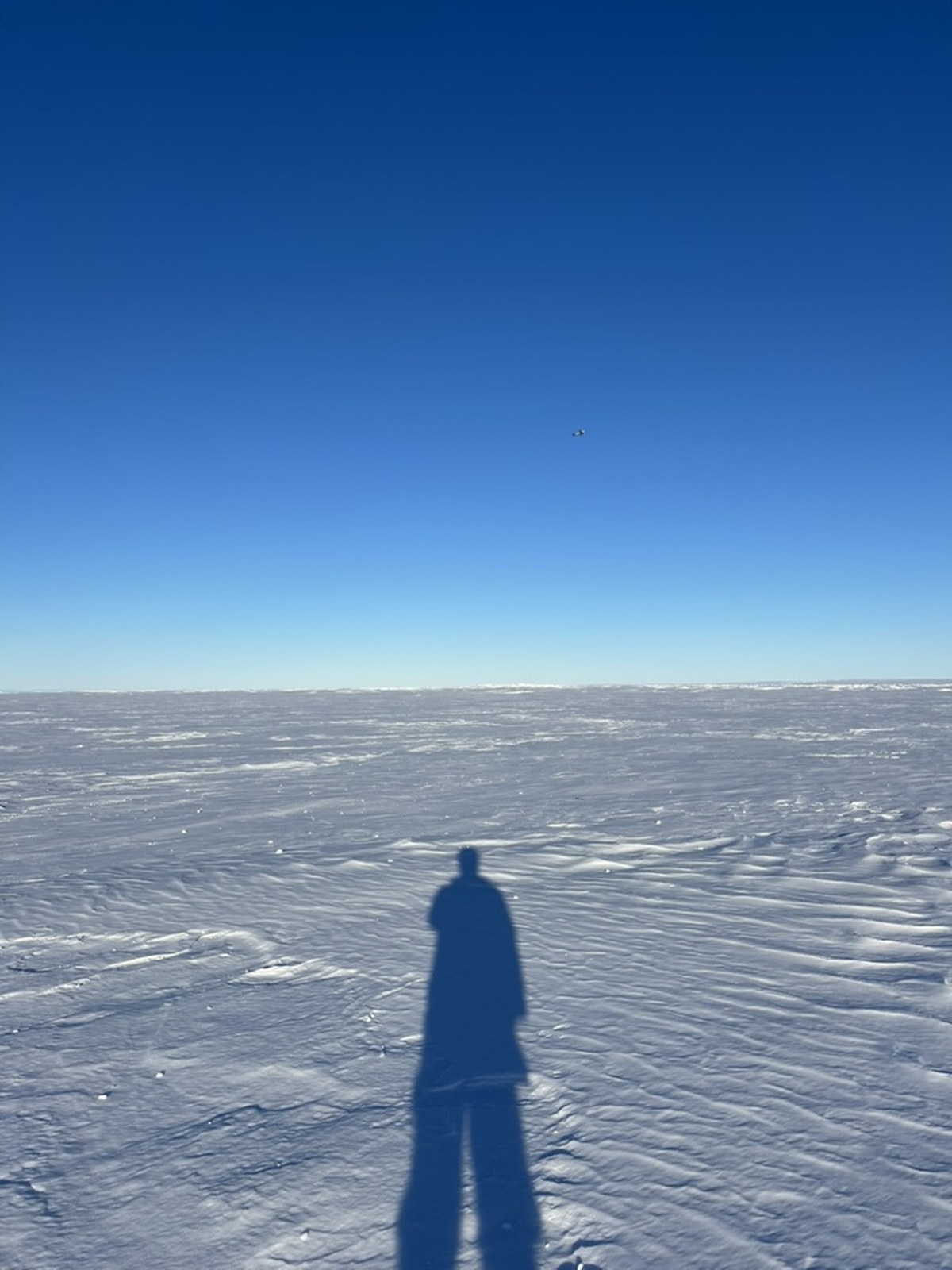 A long shadow cast on the ice at Thwaites Glacier's main trunk during the 2025–26 hot water drilling expedition, with a returning helicopter on the horizon.